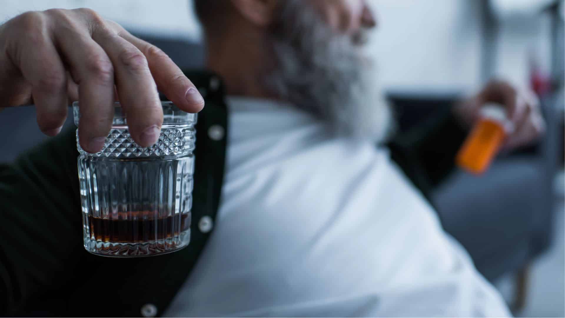 senior man holding glass with whiskey and bottle with medication at home