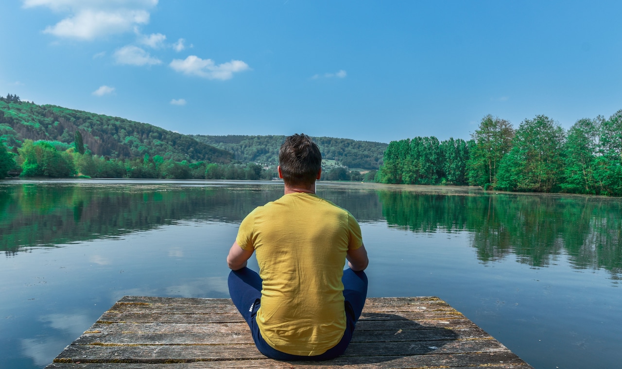 Man meditating on a wooden dock beside a calm lake under a clear sky